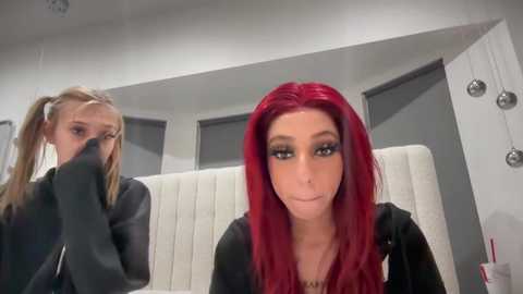 Media: Video of two women with long red hair in a modern bathroom with white walls, a gray sink, and a white toilet. The woman in the foreground is smiling, while the one in the background is covering her mouth.