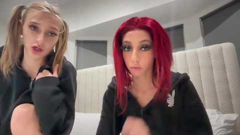 Media: Video of two young women with pale skin and red hair, wearing black Playboy bunny hoodies, sitting on a white bed in a modern, minimalist bedroom.