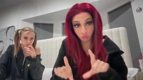 Media: Video of a red-haired woman with long hair, wearing a black hoodie, flipping the bird, standing in a modern, minimalist bedroom with white walls and a white bed. A blonde woman in a black jacket watches from the background.