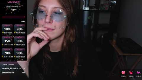 Media: Video of a young woman with long brown hair, wearing glasses, and a black shirt, with a dimly lit background of a messy kitchen, featuring a counter and appliances.