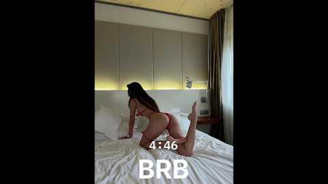 Media: Video of a woman with long brown hair, nude, on all fours on a bed, facing away. Modern, minimalist room with white bedding, gray walls, and a bedside table. Time stamp: 4:26.