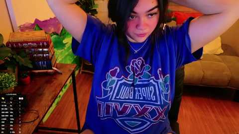 Media: Video of a young woman with medium skin tone and long black hair, wearing a blue t-shirt with \"LIVE\" and \"MUSIC\" in white and pink, seated indoors with potted plants, a wicker basket, and a couch in the background.