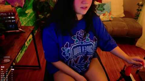 Media: Video of a woman with long black hair, wearing a blue \"Our Florida\" t-shirt, sitting on a wooden floor, playing a red controller. Background includes a green plant, a brown sofa, and a colorful blanket.