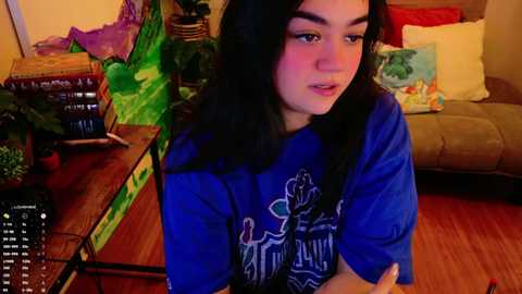 Media: Video of a young woman with long black hair, wearing a blue t-shirt, sitting on a wooden floor, surrounded by green plants, books, and a cozy living room.