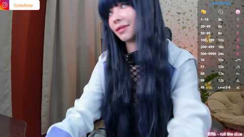 Media: A video of a young woman with long, straight, dark blue hair and pale skin, wearing a light blue jacket over a black fishnet top, standing in a room with beige curtains.