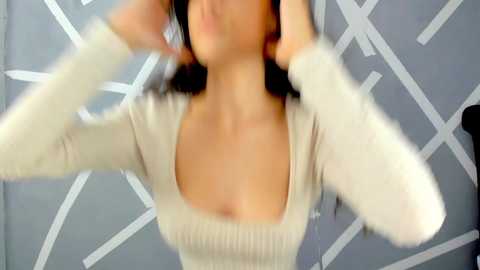 Media: Video of a woman with medium-length dark hair, wearing a cream-colored, long-sleeve, ribbed-knit top with a square neckline, adjusting her hair in front of a geometric-patterned gray wall.