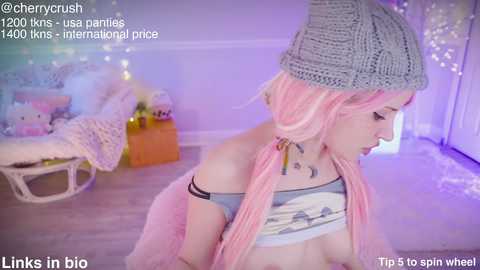 Media: Video of a young woman with pink hair, wearing a gray knitted hat and crop top, sitting in a cozy, pastel-colored room.