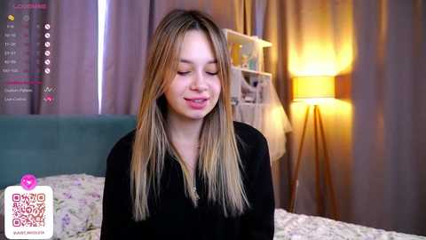 Media: Video of a young, smiling Caucasian girl with long, straight blonde hair, wearing a black shirt, in a cozy bedroom with a teal headboard, a white lamp, and a floral bedspread.