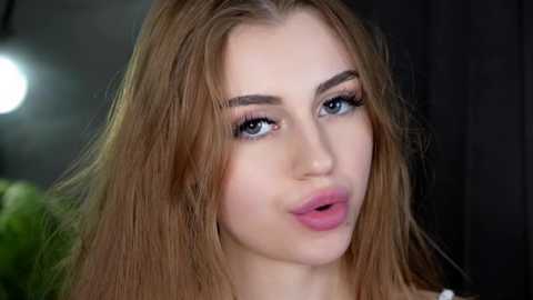 Media: Video of a young Caucasian woman with fair skin, long straight light brown hair, and blue eyes. She has full lips with pink lipstick, and she's looking directly at the camera with a neutral expression.