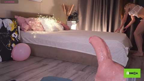 Media: Video of a cozy bedroom with a man's erect penis in the foreground, two women in lingerie on a bed with fairy lights, pink balloons, and a wooden floor.