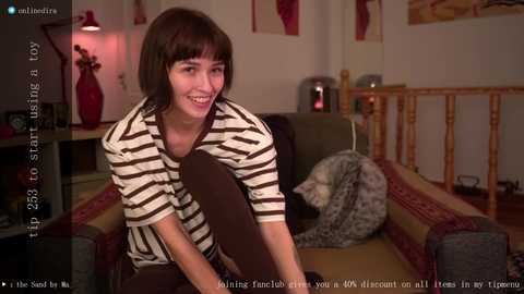 Media: Video of a woman with short dark hair in a striped shirt, sitting on a couch in a cozy living room with a wooden railing and red wall art.