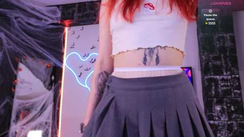 Media: Video of a slim, fair-skinned woman with long red hair, wearing a white crop top and gray pleated skirt. Background includes a cityscape print, a glowing neon heart, and a spiderweb.