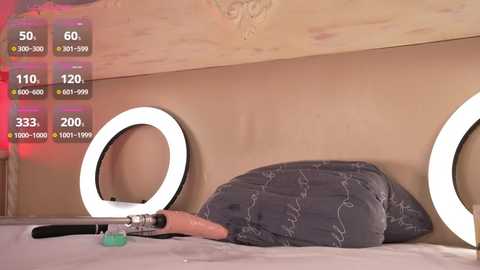 Media: Video of a bedroom with a beige wall featuring circular, glowing white light fixtures. A grey pillow with a floral pattern rests against the wall, and a pink and black hairbrush lies on a bed.