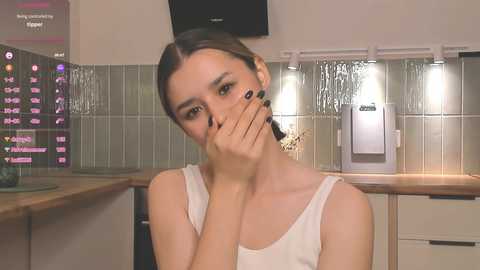 Media: A video of a young woman with fair skin and brown hair, wearing a white tank top, covering her mouth in a kitchen with tiled backsplash, countertop, and illuminated stove.