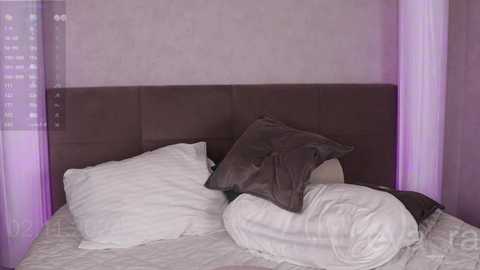 Media: A video of a modern bedroom featuring a plush brown headboard, white pillows, and a white duvet, partially covered by a brown blanket. The walls are adorned with a subtle, light purple pattern.