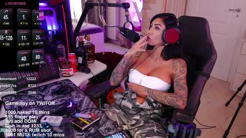 Media: Video of a tattooed woman with long black hair, wearing a white bandeau top and camo pants, sitting in a gaming chair, holding a microphone, in a cluttered room with gaming equipment.