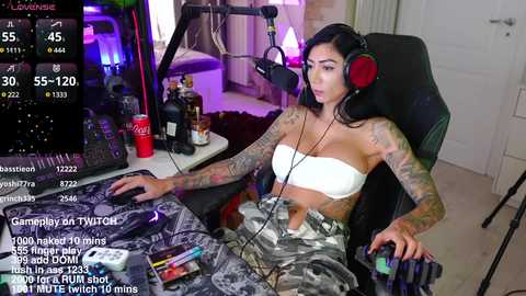 Media: Video of a tattooed woman with black hair in headphones, seated at a desk with gaming gear, drinking Coke Zero, in a dimly lit room.