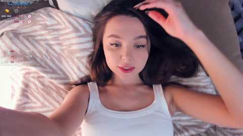 Media: A video of an Asian woman with long black hair, lying on a bed with patterned bedding, wearing a white tank top, and smiling.