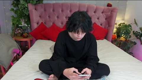 Media: Video of a young man with short, dark hair, sitting cross-legged on a bed with a pink, tufted headboard, wearing black clothes, holding a smartphone.