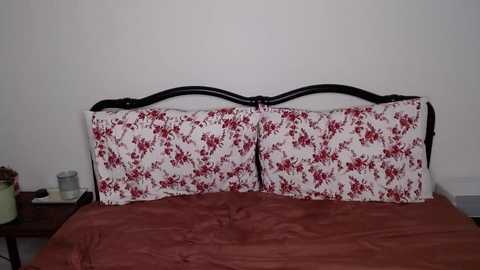 Media: A video of a neatly made bed with a dark wooden headboard, featuring two matching pillows with a white floral pattern and red accents. The bedspread is a rich maroon color, and a small green potted plant sits on the bedside table.