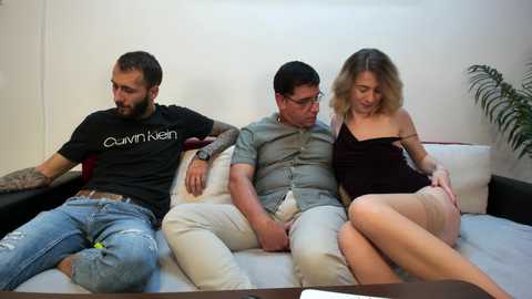 Media: Video of three young adults, two men and a woman, sitting on a beige sofa in a living room. The woman is dressed in a black spaghetti-strap top, revealing her cleavage, and the men wear casual clothing.