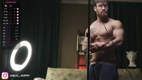 Media: Video of a shirtless, muscular man with tattoos and glasses, wearing headphones, standing in a dimly lit room with a green curtain and a lit ring light.