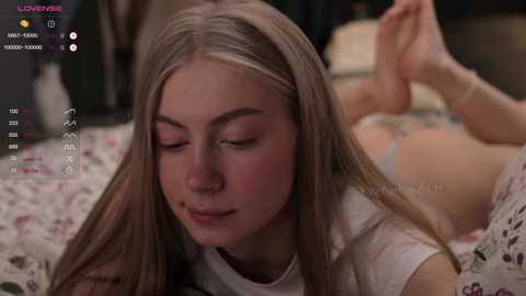 Media: Video of a young Caucasian woman with light skin and long blonde hair, lying on a floral-patterned bed, wearing a white top and white underwear, eyes closed, looking serene.