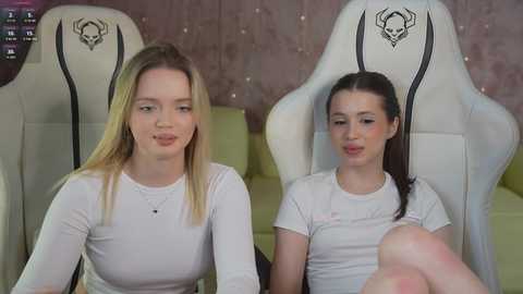 Media: Video of two young women, one blonde and one brunette, wearing white shirts, seated in white gaming chairs with black skull logos, in a room with pink wallpaper and a green couch.