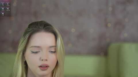 Media: A video of a young, light-skinned blonde woman with closed eyes, sitting on a green couch in a softly lit room with a textured, muted pink wall.