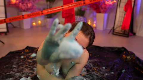 Media: Video of a nude woman with wet hair, covered in blue paint, lying on a black tarp in a dimly lit room with red and orange lights, and caution tape in the background.