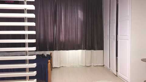 Media: Video of a small, sparsely furnished room with a bed against a dark, vertical striped curtain. A white door is on the right, and a white blind covers part of the left wall.