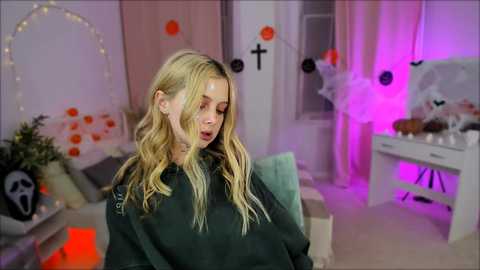 Media: Video of a blonde woman with wavy hair in a dark hoodie, standing in a Halloween-themed room with orange and purple lights, decorations, and a white desk.