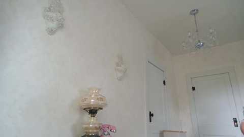 Media: Video of a minimalist hallway with cream walls, featuring a chandelier and decorative sconces, a vintage lamp, and a pink flower arrangement.