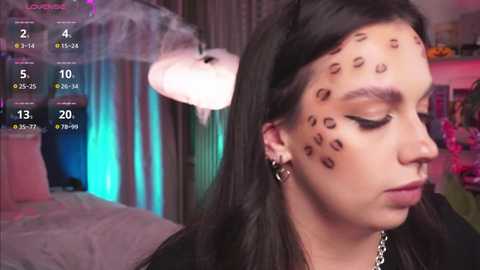 Media: Video of a young woman with long black hair and leopard print face paint, wearing a black top and silver earrings, indoors with a bed and colorful room in the background.