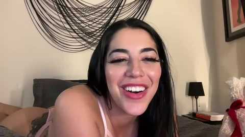 Media: Video of a smiling, fair-skinned woman with long black hair, wearing a pink tank top, in a modern bedroom with a black woven wall hanging and a black lamp on a nightstand.