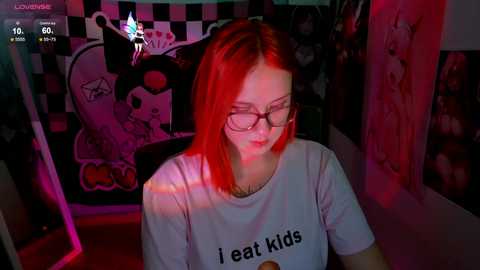 Media: Video of a young, fair-skinned woman with straight red hair, wearing glasses and a \"I eat kids\" T-shirt, sitting at a gaming chair in a dimly lit, decorated room with anime posters.