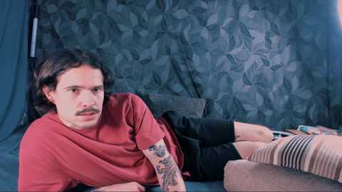 Media: Video of a man with shoulder-length brown hair and a mustache, wearing a red T-shirt, black shorts, and striped socks, lying on a bed with a teal patterned backdrop.