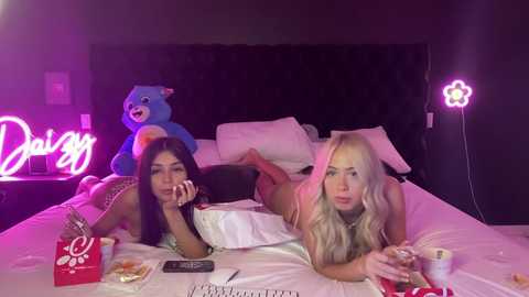 Media: Video of two women lying on a bed, wearing lingerie, surrounded by neon lights and stuffed toys in a dimly-lit room.