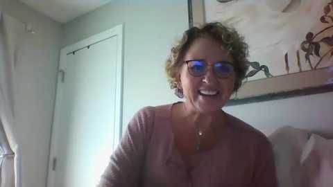 Media: Video of a smiling older woman with curly blonde hair, wearing glasses and a pink cardigan, in a softly lit room with beige walls, a framed painting, and a white door.