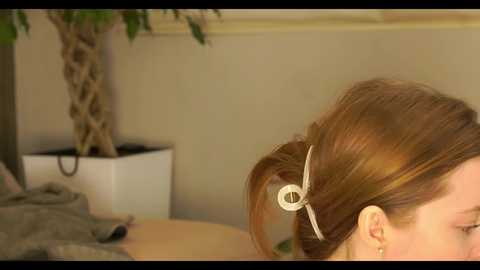 Media: Video of a woman with light brown hair tied in a bun with a white hair tie, partially visible. Background includes a beige wall, a palm tree in a white planter, and a blurred bed with grey bedding.