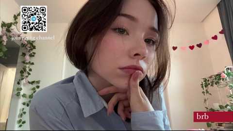 Media: A video of a young woman with fair skin, straight brown hair, and light makeup, wearing a light blue shirt, in a cozy, floral-decorated room.