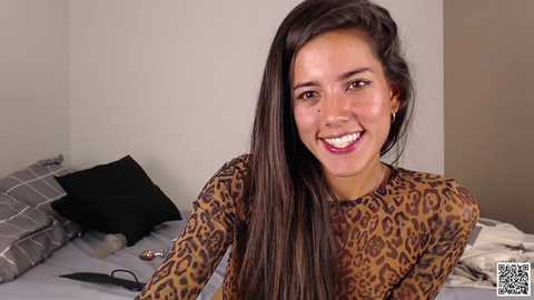 Media: Video of a young, smiling Latina woman with long, dark hair, wearing a leopard print top, sitting on a bed with grey and black plaid pillows.