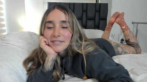 Media: A video of a tattooed woman with long, wavy hair lying on her stomach on a bed, wearing patterned socks and a black shirt.