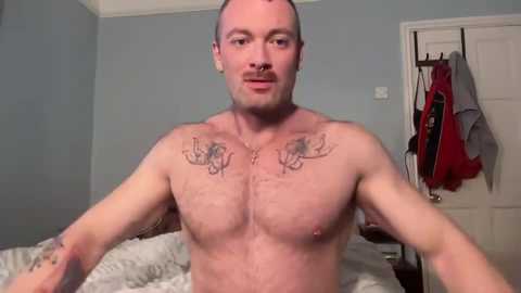 Media: Video of a shirtless, muscular, bald Caucasian man with tattoos of angel wings on his chest, standing in a bedroom with a white bed, blue walls, and hanging clothes.