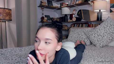 Media: A video of a young Asian woman with dark hair in a bun, lying on a bed, holding a phone, in a cozy room with bookshelves and patterned pillows.