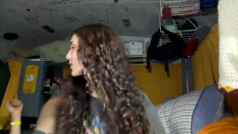 Media: Video of a young woman with long, curly brown hair, standing in a cluttered bedroom with yellow walls, a hanging black bra, and a bed with striped sheets.