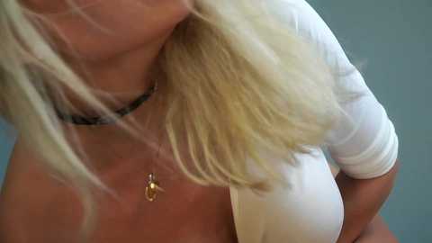 Media: A close-up video of a blonde woman with shoulder-length hair, wearing a white top, and a black choker necklace with a pendant. The background is a blurred, light blue wall.