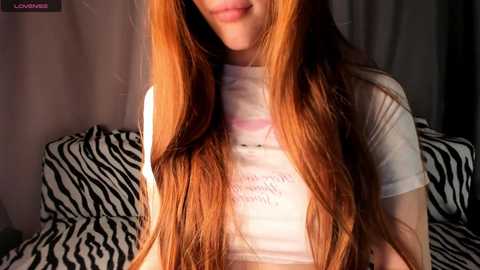 Media: Video of a young woman with long, straight, auburn hair, wearing a white t-shirt with pink text, smiling, sitting on a black-and-white zebra print couch in a dimly lit room.