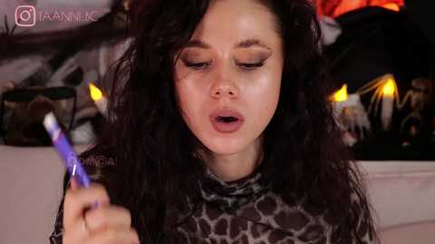 Media: A video of a woman with curly black hair and fair skin, wearing a leopard-print top, holding a purple lipstick, with candles and Halloween decor in the background.