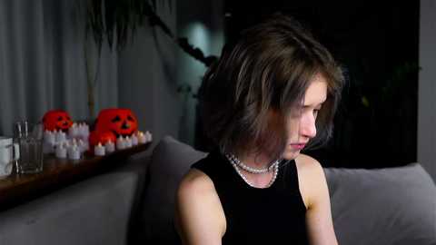 Media: A video of a woman with shoulder-length, wavy brown hair in a black dress, seated on a grey sofa. She wears a pearl necklace. The background features a wooden shelf with white candles and a red Jack-o'-lantern. The room has dim lighting and a dark, Halloween-themed ambiance.